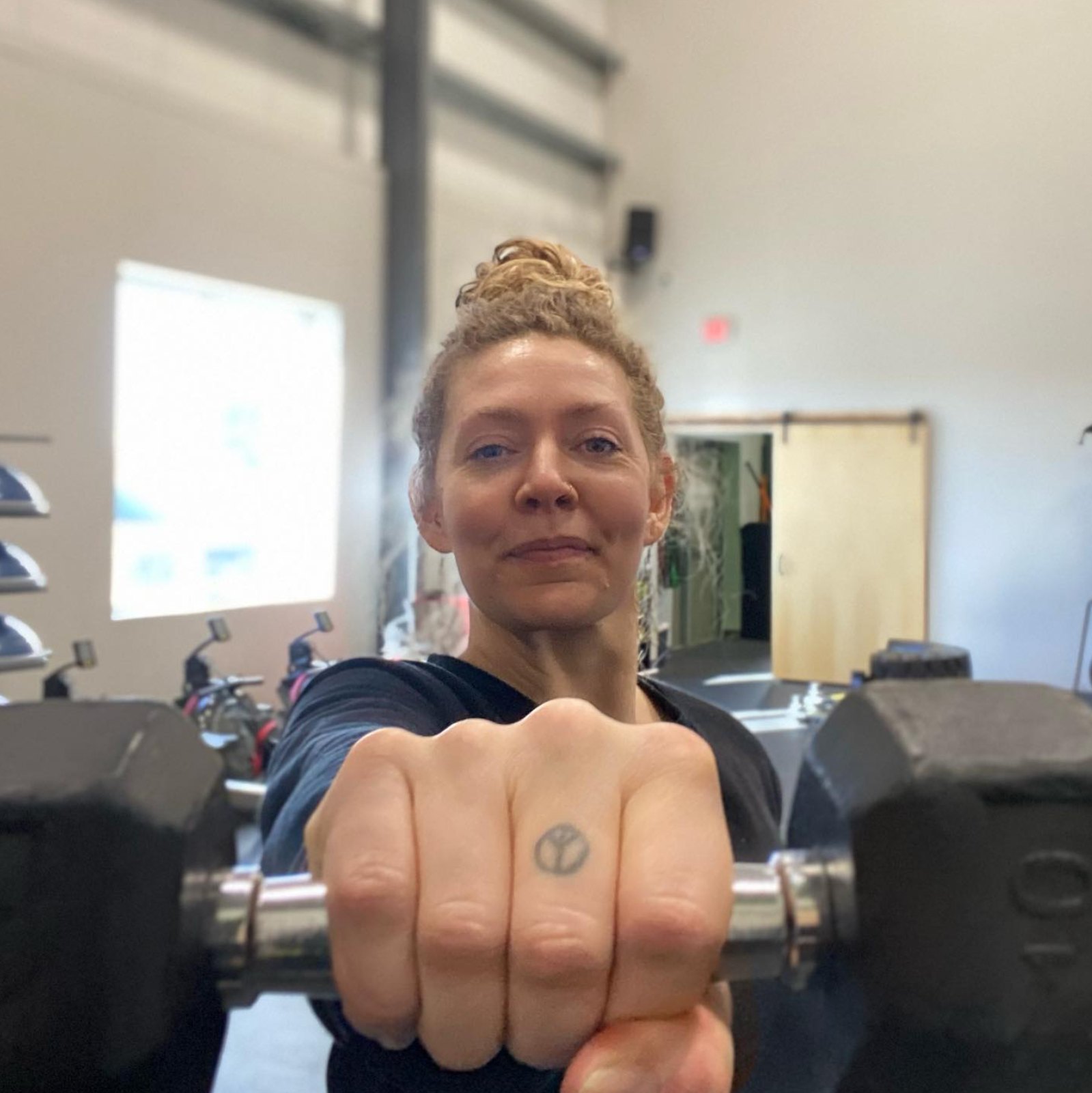 Rachel Hamilton holding a dumbbell toward the camera, knuckle tattoo visible, at the gym